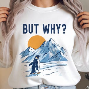 Könnte beinhalten: Weißes Sweatshirt mit dem Text "BUT WHY?" über einer Grafik eines Pinguins, der vor einer Bergkette und einer gelben Sonne steht. Der Pinguin ist schwarz und weiß.