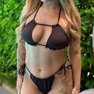 Könnte beinhalten: Schwarzes Bikini-Set mit Neckholder-Top und Schlüsselloch-Design, mit Rüschenbesatz. Die passenden Unterteile haben Bindebänder an den Seiten. Die Bademode ist aus einem glatten, dunklen Stoff gefertigt.
