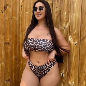 Könnte beinhalten: Ein zweiteiliger Bikini mit Leopardenmuster. Das trägerlose Oberteil und der hoch taillierte Slip sind braun und schwarz. Die Person trägt eine Sonnenbrille und eine Halskette. Der Hintergrund ist eine Holzwand.
