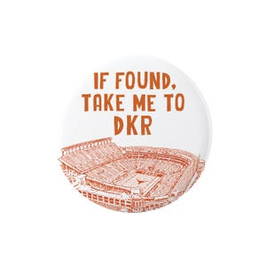 May include: A round white pinback button featuring orange text that reads IF FOUND, TAKE ME TO DKR. Below the text is an orange line drawing of a large football stadium with the word TEXAS visible on the field and a longhorn logo at midfield.