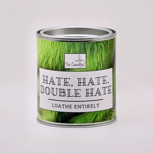 May include: A round tin candle with a green, textured surface. The label displays the text "HATE, HATE, DOUBLE HATE" and "LOATHE ENTIRELY" in black. The top is silver, and the brand logo "Tin Candles" is also visible.