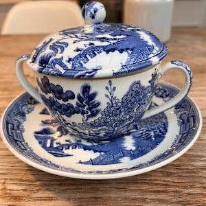 Puede incluir: Juego de té de porcelana azul y blanca con taza, platillo y azucarero con tapa. El juego presenta un detallado diseño de paisaje. La taza tiene dos asas y la tapa un pequeño pomo esférico. El juego está sobre una superficie de madera.