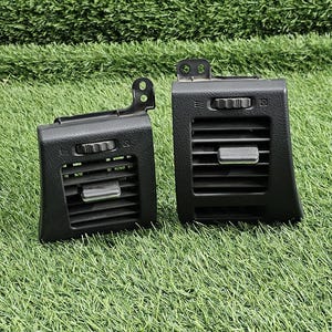 May include: Two black plastic automotive air vent assemblies featuring horizontal slats, a central adjustment tab, and a textured finish. Each unit includes a mounting bracket on the top edge and integrated controls for airflow direction and intensity.