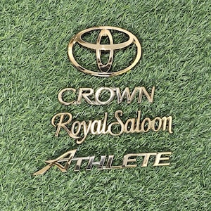 Puede incluir: Emblemas dorados de Toyota Crown Royal Saloon Athlete sobre un fondo verde. El logotipo de Toyota está en la parte superior, seguido del texto "CROWN Royal Saloon" y "ATHLETE".