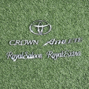 Puede incluir: Un emblema y texto plateados de Toyota sobre un fondo verde. El texto incluye "CROWN Athlete", "Royal Saloon" y "Royal Extra". El logotipo de Toyota está en la parte superior central.