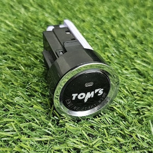 Op de afbeelding: Een zwarte en chromen motorstartknop met het TOM'S-logo in witte tekst. De ronde knop heeft een glanzend zwart midden met de woorden ENGINE START in kleine witte letters gedrukt langs de onderste rand van de ring.