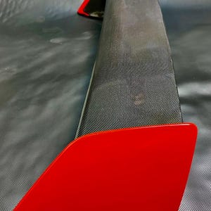 May include: A red and black car spoiler. The spoiler has a glossy red finish on the edges and a textured black surface in the center. The design is aerodynamic, suggesting a performance-oriented automotive accessory.