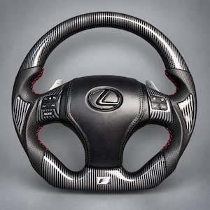 Puede incluir: Un volante Lexus con acabado en fibra de carbono negra en las secciones superior e inferior, agarres laterales de cuero perforado negro con costuras rojas, botones de control integrados, levas de cambio plateadas y un emblema F en la base.