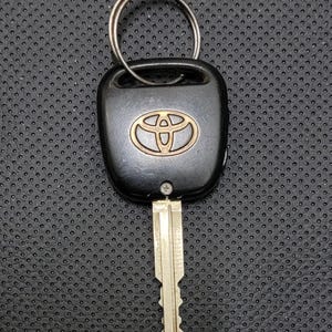 Könnte beinhalten: Schwarzer Toyota Autoschlüssel mit einem silbernen Schlüsselring. Der Schlüssel hat ein schwarzes Kunststoffgehäuse mit dem Toyota-Logo in Gold. Die Schlüsselklinge ist silberfarben und hat eine geriffelte Kante.