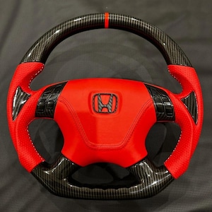 Puede incluir: Un volante de coche personalizado con centro y detalles en cuero rojo, con molduras de fibra de carbono. El volante tiene un logotipo negro de Honda y una raya roja en la parte superior. Costuras blancas.