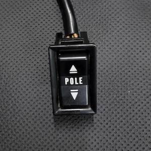 Puede incluir: Un interruptor rectangular negro con la palabra "POLE" en texto blanco. El interruptor tiene un triángulo que apunta hacia arriba encima de la palabra y un triángulo que apunta hacia abajo debajo. Un cable negro se extiende desde la parte superior del interruptor.
