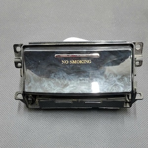 Może przedstawiać: Czarny popielniczka samochodowa ze złotym napisem "NO SMOKING". Popielniczka ma prostokątny kształt z błyszczącą, odblaskową powierzchnią. Posiada wsporniki montażowe po każdej stronie.