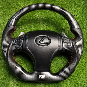 Puede incluir: Un volante de fibra de carbono negro y plateado con costuras rojas. El volante presenta el logotipo de Lexus y el emblema F Sport. El volante tiene botones para el modo y la pantalla.