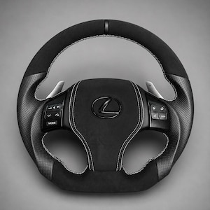 Puede incluir: Un volante Lexus negro que combina texturas de cuero liso y perforado, con costuras blancas en contraste, una marca central superior, botones de control integrados en los radios horizontales y levas de cambio de marchas plateadas.