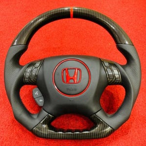 Könnte beinhalten: Schwarzes Lenkrad aus Kohlefaser mit rotem Honda-Emblem in der Mitte. Das Lenkrad hat perforierte Ledergriffe und integrierte Bedienelemente. Ein roter Streifen akzentuiert die Oberseite des Lenkrads.