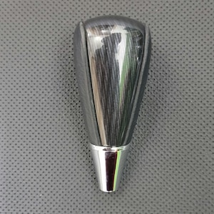 Puede incluir: Un pomo de palanca de cambios negro y plateado con acabado brillante. La parte superior es negra con un patrón de vetas de madera y la base es plateada. El pomo está diseñado para la transmisión de un vehículo.