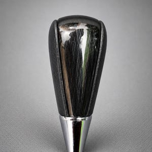 Peut inclure: Un pommeau de levier de vitesse doté d'un panneau central au motif de grain de bois sombre et vertical, recouvert d'une finition brillante. Les côtés sont gainés de matière noire et la base est un col en métal argenté poli et conique.
