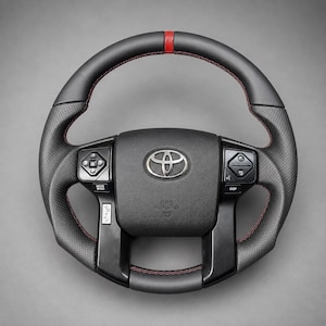 Könnte beinhalten: Ein schwarzes Lederlenkrad mit einem roten Mittelstreifen oben, perforierten Ledergriffen an den Seiten und roten Kontrastnähten. Die mittlere Nabe zeigt ein silbernes Toyota-Emblem und integrierte schwarze Bedientasten auf beiden Seiten.