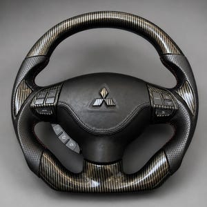 Pode incluir: Um volante preto com acabamento em fibra de carbono nas seções superior e inferior, com pegas laterais em couro preto perfurado e costuras vermelhas. O centro exibe um logotipo prateado da Mitsubishi e botões de controle integrados.