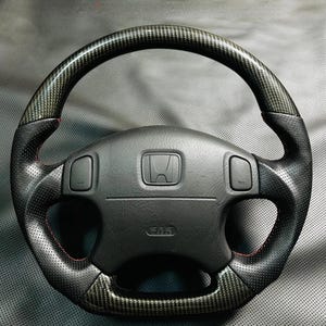 Könnte beinhalten: Ein schwarzes Lenkrad mit Carbonfaser-Akzenten. Das Lenkrad hat einen perforierten Ledergriff mit roten Nähten und ein Honda-Emblem in der Mitte. Der Airbag ist mit "SRS" beschriftet.