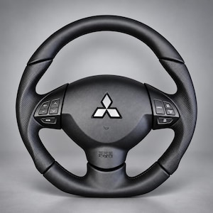 May include: A black Mitsubishi steering wheel featuring a central silver three-diamond logo, perforated leather side grips, and integrated control buttons on both the left and right spokes. The bottom spoke includes an embossed SRS AIRBAG label.