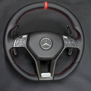 May include: Black leather steering wheel with red stitching and a carbon fibre centre. Features a red stripe at the top and paddle shifters. The centre has the Mercedes-Benz logo and an AMG emblem.