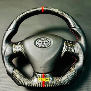 May include: A black and carbon fibre steering wheel with red stitching and accents. The wheel features a Toyota logo and the letters "TRD" in yellow. The top of the wheel has a red stripe.