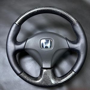 Puede incluir: Un volante negro y de fibra de carbono con el logotipo de Honda en el centro. El volante tiene una parte superior e inferior de fibra de carbono con laterales de cuero negro. El airbag está etiquetado como "SRS AIRBAG".