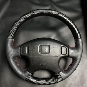 May include: Black steering wheel with carbon fiber accents and red stitching. The wheel features a perforated leather grip and a center emblem. The design includes integrated buttons and a carbon fiber top.
