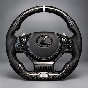 Puede incluir: Un volante Lexus negro con acabado de fibra de carbono en las secciones superior e inferior, agarres laterales de cuero negro perforado, una franja central blanca en la parte superior y un emblema F Sport plateado en la parte inferior con botones.