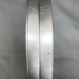 Op de afbeelding: Twee grijze auto-sierlijsten, waarschijnlijk voor een autodeur of carrosseriepaneel. De gebogen stukken hebben een metallic glans, wat wijst op een modern ontwerp. Ze zijn verticaal geplaatst tegen een getextureerde grijze achtergrond.