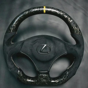 May include: A black and carbon fibre steering wheel with a yellow stripe at the top. The wheel has a black suede centre with the Lexus logo and white stitching. The carbon fibre sections have a marbled appearance.