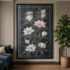 May include: A framed stained glass panel featuring a magnolia branch with white and pale pink blossoms and green leaves against a dark blue background. The artwork is in a black frame and is displayed in a room with a plant.