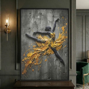 May include: A framed artwork featuring a ballerina in mid-air, rendered in black and gold against a textured gray background. The ballerina's dress is a splash of gold paint, creating a dynamic and artistic effect. The artwork is displayed in a dark room.