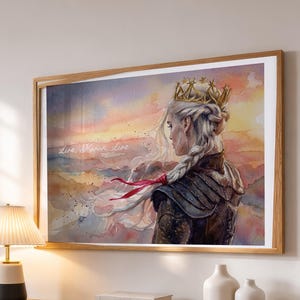 May include: Watercolor print of a woman with long, white braided hair, wearing a gold crown and armor, set against a sunset backdrop. The artwork is framed in a light brown wooden frame. The text "Live, Warm, Live" is visible.