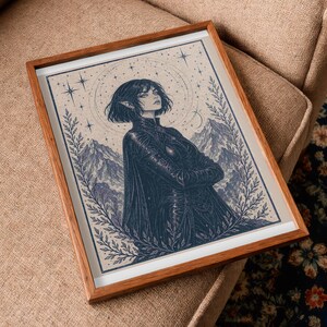 May include: Framed art print featuring a detailed illustration of a person with short hair, arms crossed, and a dark outfit. The artwork is set against a backdrop of mountains, stars, and a celestial design, framed in a wooden frame.