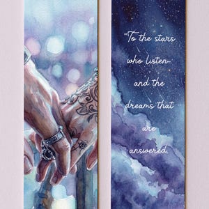 May include: Two watercolor bookmarks. One features hands holding each other, with intricate tattoos and rings. The other has a starry night sky with the quote, "To the stars who listen and the dreams that are answered."