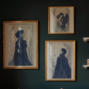 May include: Three framed art prints with gold frames hang on a dark green wall. The prints feature illustrations of figures in dark blue, with celestial and botanical elements. One print depicts a couple embracing, another a figure looking up, and the third a woman.