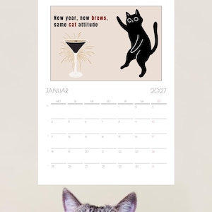 May include: A 2027 wall calendar for the month of January featuring a black cat illustration, a martini glass, and the text New year, new brews, same cat attitude. The calendar grid displays the days of the month with the header JANUAR 2027.