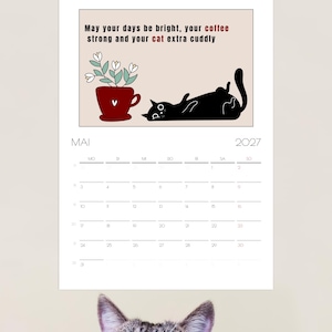 May include: A wall calendar for May 2027 featuring a black cat lying on its back next to a red mug with white flowers. The text reads: May your days be bright, your coffee strong and your cat extra cuddly. A tabby cat head is visible at the bottom.