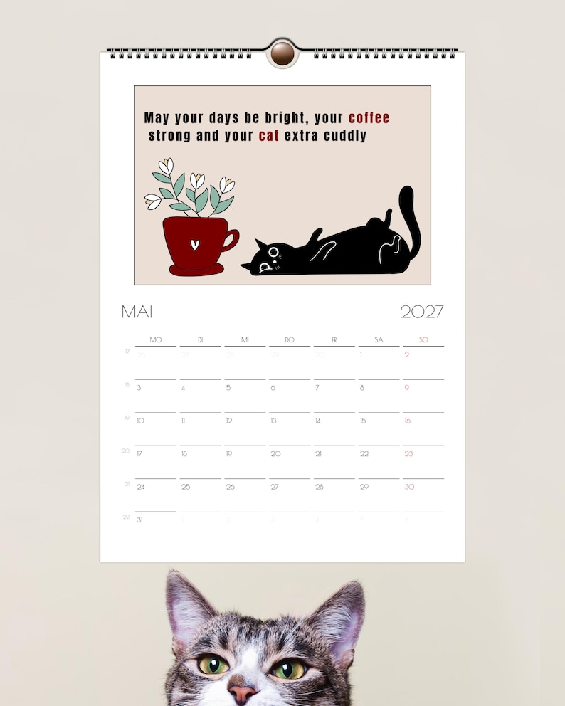 May include: A wall calendar for May 2027 featuring a black cat lying on its back next to a red mug with white flowers. The text reads: May your days be bright, your coffee strong and your cat extra cuddly. A tabby cat head is visible at the bottom.