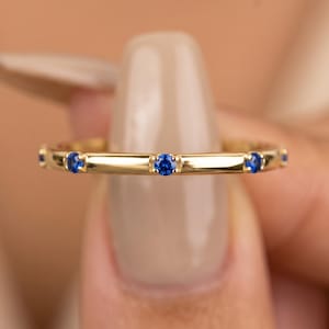 Op de afbeelding: Een dunne gouden ring met een reeks kleine, ronde, briljant geslepen blauwe edelstenen die met regelmatige tussenpozen langs de buitenomtrek van de metalen band zijn gezet, met een gepolijste afwerking en een verfijnde constructie.
