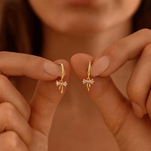 Puede incluir: Un par de pendientes de aro dorados, cada uno con un pequeño diseño de lazo con detalles claros y brillantes. Los pendientes se muestran de cerca, mostrando sus delicados detalles y su estilo elegante.