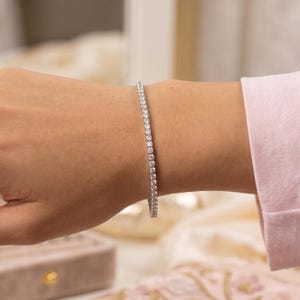 May include: A silver-toned tennis bracelet featuring a continuous row of small, round, clear, faceted stones set in a metallic frame. The bracelet encircles the wrist, showcasing a uniform design with sparkling, reflective elements throughout the piece.