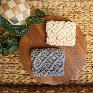 May include: Two handmade crochet coasters. One is cream-colored, the other is gray. Both coasters have a textured, diagonal pattern. They are displayed on a round, wooden tray, with a woven background and decorative elements.