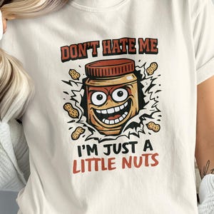 May include: Cream-colored t-shirt featuring a cartoon peanut butter jar with a smiling face and the text "DON'T HATE ME I'M JUST A LITTLE NUTS". The design includes peanuts and a brown jar lid.