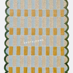 May include: A rectangular rug with a checkered pattern in light blue, tan, and mustard yellow. The rug has a scalloped green border and the text "KNOT & LOOMS" is visible. The rug's design features vertical and horizontal rectangles.