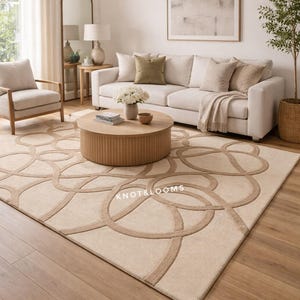 Puede incluir: Alfombra beige con un patrón circular repetitivo en un tono beige más claro. La alfombra está en un salón con un sofá blanco, una mesa de centro de madera y un árbol en una cesta. El texto "KNOT & LOOMS" es visible.