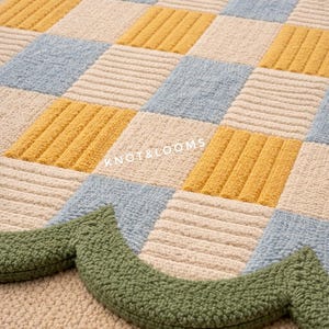 May include: Close-up of a tufted rug with a checkerboard pattern in yellow, light blue, and cream colours. A green, scalloped border accents the bottom edge. The text "KNOT&LOOMS" is visible.