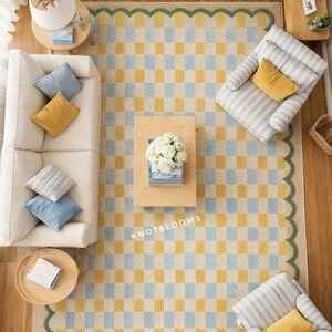 May include: An aerial view of a living room featuring a large rug with a yellow and pale blue chequered pattern and a green scalloped border. A white sofa, striped armchairs, and wooden tables are arranged around the rug. Cushions in blue and yellow.